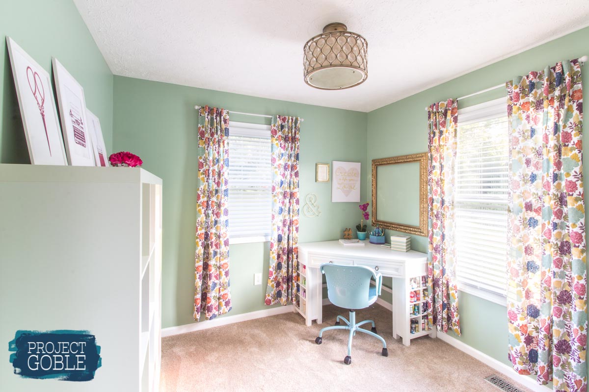 Mint Green home craft room office with white corner desk, white Ikea shelf, gold diamond light fixture, and colorful pink and purple floral curtains.